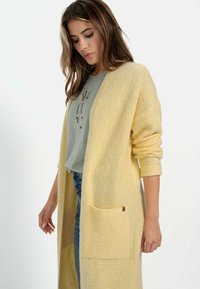 Lange gele cardigan van zacht, textuur gebreid stof, met een voorvak en opgerolde mouwen, gelaagd over een grijs shirt.