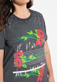 Kulkeblød grå t-shirt med røde blomster designs og fed hvid tekst "VÆR GLAD" med grønne accenter. Pyntet med sølvnitter på skuldrene.