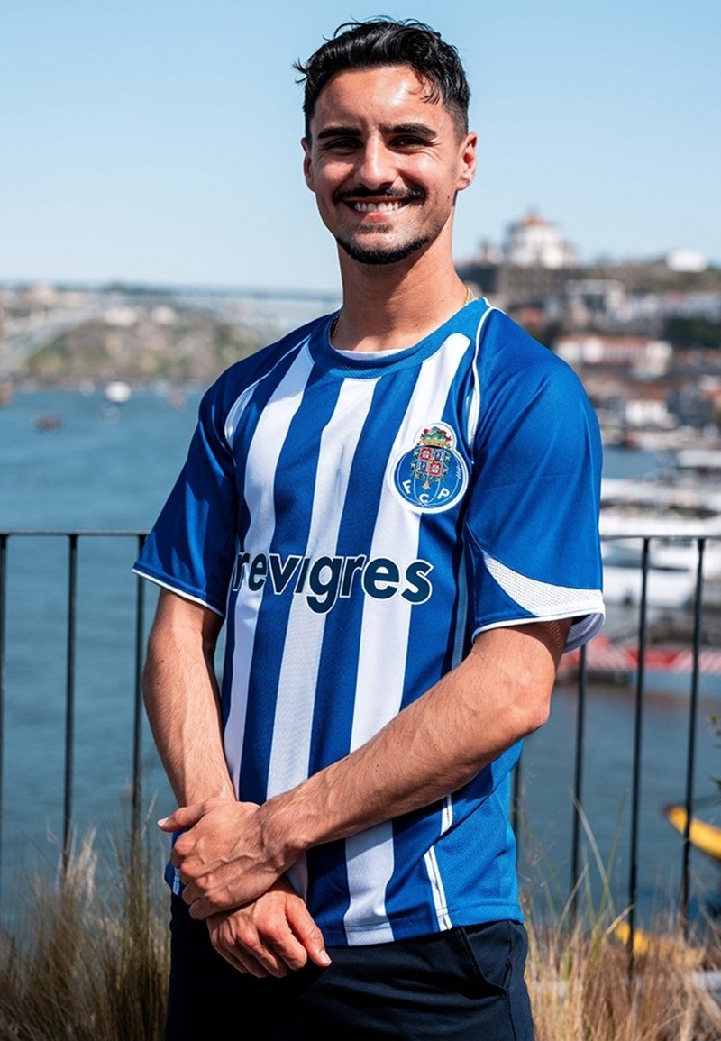 FC Porto thuisshirt met verticale blauwe en witte strepen, ronde kraag, logo op de borst, gedragen door een persoon buiten met water op de achtergrond.