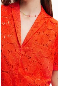 Camicia rossa in pizzo con motivi floreali, maniche corte e colletto a scollo. I bottoni sono visibili e c'è una tasca sul lato sinistro.