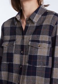 Chemise en flanelle avec un motif à carreaux bleu marine, marron et blanc, dotée d'un col classique, de deux poches poitrine et de fermetures à boutons.