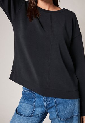 Femme portant un haut noir ample à manches longues avec des coutures visibles et un jean bleu avec de grandes poches avant.