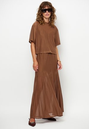 Vrouw met krullend haar die een oversized bruin gaastop en lange bijpassende rok draagt, donkere zonnebril, bruine puntige schoenen en gouden sieraden.