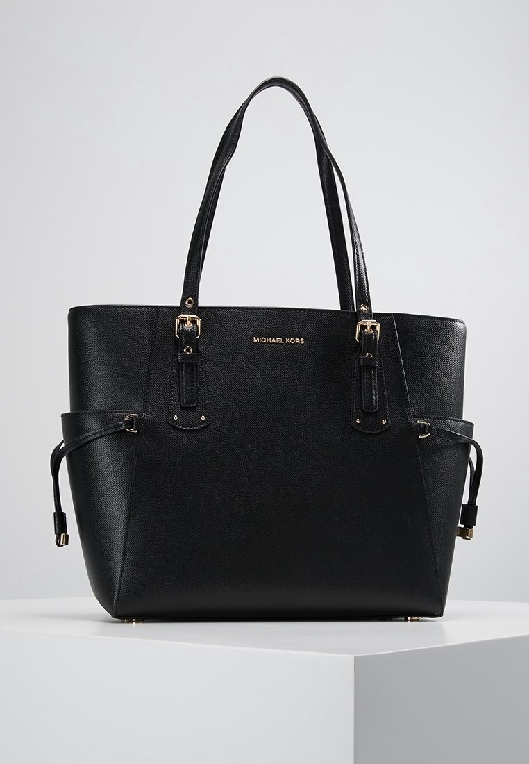 Sac cabas en cuir noir Michael Kors avec double poignée, ferrures dorées et détails à cordon latéraux sur un socle blanc.