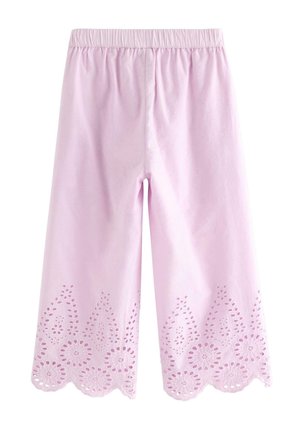 Smilende ung pige iført en bredskygget stråhat, hvid bluse med hulmønster, lyserøde bukser med hulmønster og cremefarvede ballet flats mod en ensfarvet baggrund.