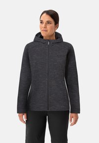 Vrouw in een donkergrijze ritsjack met capuchon en subtiele textuurpatroon, samen met zwarte broek, staande voor een effen lichte achtergrond.