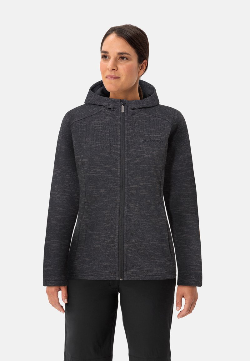 Vrouw in een donkergrijze ritsjack met capuchon en subtiele textuurpatroon, samen met zwarte broek, staande voor een effen lichte achtergrond.