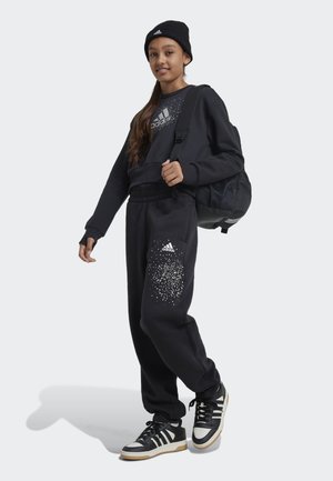 Sudadera negra con logo, joggers a juego con un diseño moteado y zapatos negros con acentos blancos; complementado con una mochila negra.