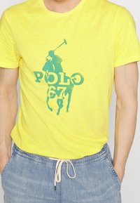 Keltainen puuvillapaita, jossa on sininen polo-pelaajan graafinen kuva ja teksti "POLO 67." Lyhyet hihat, pyöreä pääntie ja pehmeä rakenne.