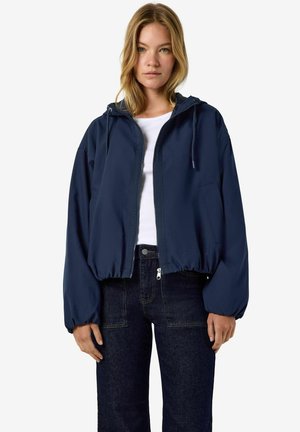 Femme portant une veste à capuche bleu marine zippée sur une chemise blanche et un jean bleu foncé, debout devant un fond blanc uni.