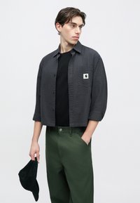 Γκρι πουκάμισο με σχέδιο και κουμπιά, με κεντημένο λογότυπο, φορεμένο πάνω από μαύρη μπλούζα και συνδυασμένο με πράσινο παντελόνι, κρατώντας μαύρο καπέλο στο χέρι.