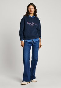 Felpa navy con testo del logo bianco e rosa, abbinata a jeans flared di jeans blu. La modella indossa una camicia col celeste sotto.