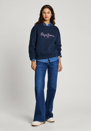 Felpa navy con testo del logo bianco e rosa, abbinata a jeans flared di jeans blu. La modella indossa una camicia col celeste sotto.