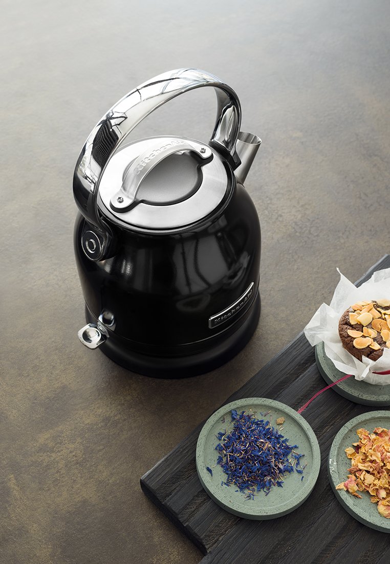 Czarny czajnik z akcentami ze stali nierdzewnej, o zaokrąglonym kształcie i ergonomicznej rączce. Zawiera dzióbek i stoi obok ozdobnych misek z składnikami do herbaty.