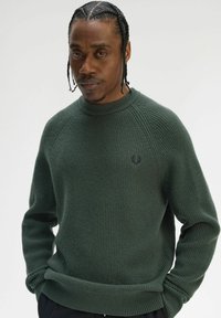 Maglione verde scuro a coste con scollo rotondo, maniche lunghe e piccolo logo sul petto. Texture liscia e vestibilità comoda.