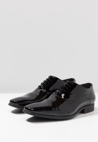 Chaussures de ville pour hommes en cuir verni noir avec un laçage fermé et des bouts capitonnés, placées côte à côte sur une surface blanche avec un fond uni.