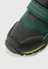 Czarno-zielony dziecięcy but z paskiem na rzep, teksturowaną górą z tkaniny, wzmocnionym noskiem i jaskrawożółtymi przeszyciami jako akcentami.