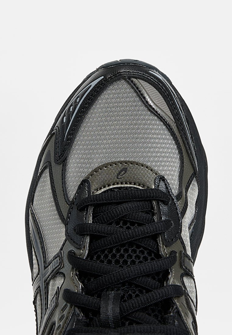 Gros plan sur une chaussure de sport noire et grise avec une tige en mesh, des coutures détaillées et des lacets noirs noués au centre.