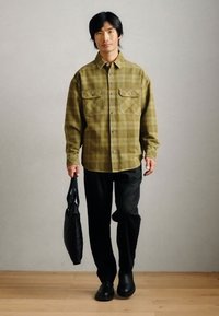 Chemise à carreaux vert olive avec deux poches poitrine, associée à un pantalon noir et des chaussures noires à enfiler ; tenant un sac noir.