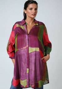 Frau in einem lockeren, langärmligen Hemdkleid mit einem bunten, abstrakten Muster in Lila, Rot und Grün, mit Händen in den Taschen, vor einem schlichten Hintergrund.
