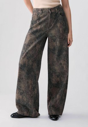 Femme portant un jean large à taille haute avec un motif abstrait foncé, un haut sans manches crème et des chaussures plates noires, debout.