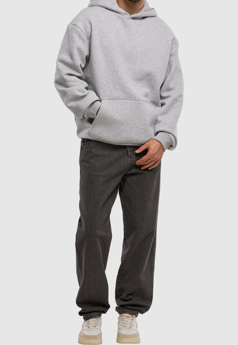 Homme portant un sweat à capuche gris clair, un jean ample gris foncé et des baskets blanches, debout devant un fond uni.