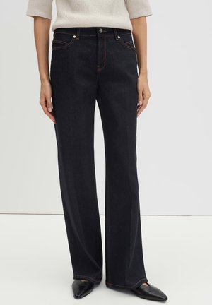 Person trägt schwarze Jeans mit hoher Taille und geradem Bein, weißes kurzärmeliges Strickoberteil und schwarze spitze Schuhe und steht vor einem einfarbigen Hintergrund.