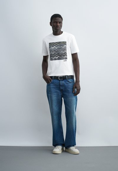 Άνδρας που φορά λευκό μπλουζάκι με στάμπα, μπλε τζιν με μαύρη ζώνη, λευκά αθλητικά παπούτσια, στέκεται μπροστά σε απλό ανοιχτογριζο φόντο.