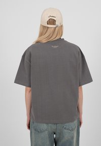 Personne aux longs cheveux blonds portant une casquette beige et un t-shirt gris surdimensionné associé à un jean bleu ample, vue de dos sur un fond gris.