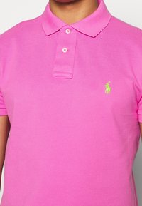 Rosa polo-skjorta i ett strukturerat tyg, med en tre-knapps knäppning och en grön logotyp i vänster bröst. Korta ärmar.