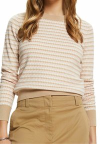 Vrouw in een beige en witte gebreide trui met lange mouwen, ingestopt in hoge taille beige broeken, met losse golvende haren zichtbaar.