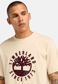 Beige bavlněné tričko s kulatým logo Timberland v barvě kaštanové, obsahující grafiku stromu a nápis "OD ROKU 1973." Krátké rukávy a kulatý výstřih.