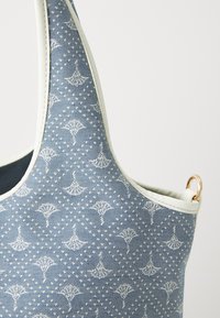 Sininen farkkutavaratykki, jossa on teksturoitu kuvio valkoisista muodoista, vaaleanbeige reuna ja kultainen akcenttirengas hihnassa.
