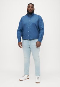 Modrá košeľa s button-up strihom vyrobená z hladkej tkaniny, spojená so svetlomodrými džínsami a bielymi teniskami s hnedými akcentmi.