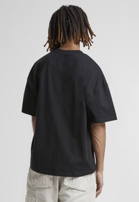 Persona con dreadlocks de longitud media, vistiendo una camiseta negra holgada y pantalones cargo de color claro, de espaldas contra un fondo simple.