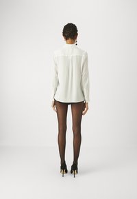 Hvid langærmet bluse med buet kant, knaplukning ved manchetterne og plissering på bagsiden, parret med gennemsigtige sorte strømpebukser og sorte hæle.