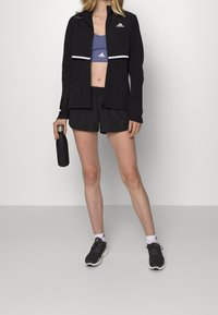 Chaqueta deportiva negra con acentos blancos, sujetador deportivo morado y pantalones cortos negros. Zapatillas negras y una botella de agua negra mate.