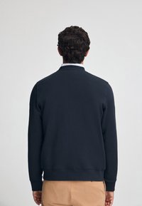 Sudadera navy con cuello redondo, puños y dobladillo acanalados. Tejido suave con una textura lisa. Llevada con pantalones beige, vista desde atrás.