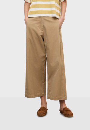 Personne portant un pantalon large beige coupé, un haut rayé beige et blanc, et des mocassins marron, les mains dans les poches.