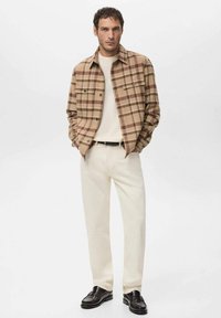 Chaqueta camisa de cuadros marrón con dos bolsillos en el pecho, suéter color crema, pantalones beige claro y mocasines marrón oscuro. Textura de tela suave.