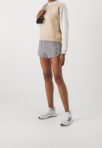 Béžová mikina s bílými rukávy, šedé šortky a bílé tenisky. Má loga Adidas a texturovaný design látky. Černé akcenty na  šortkách.