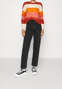 Persoon in een gestreepte oranje, roze en crèmekleurige trui, zwarte high-waisted jeans, witte sokken en zwarte lage sneakers.