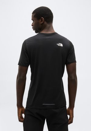 Mann, der mit dem Rücken zur Kamera steht, trägt ein schwarzes Kurzarmshirt von The North Face mit Logo auf dem oberen Rücken vor einem einfarbigen hellen Hintergrund.