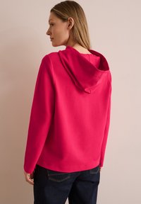 Sudadera fucsia con un corte relajado y un bolsillo canguro frontal, que presenta una capucha texturizada y puños acanalados. Combinada con unos jeans de mezclilla oscura.