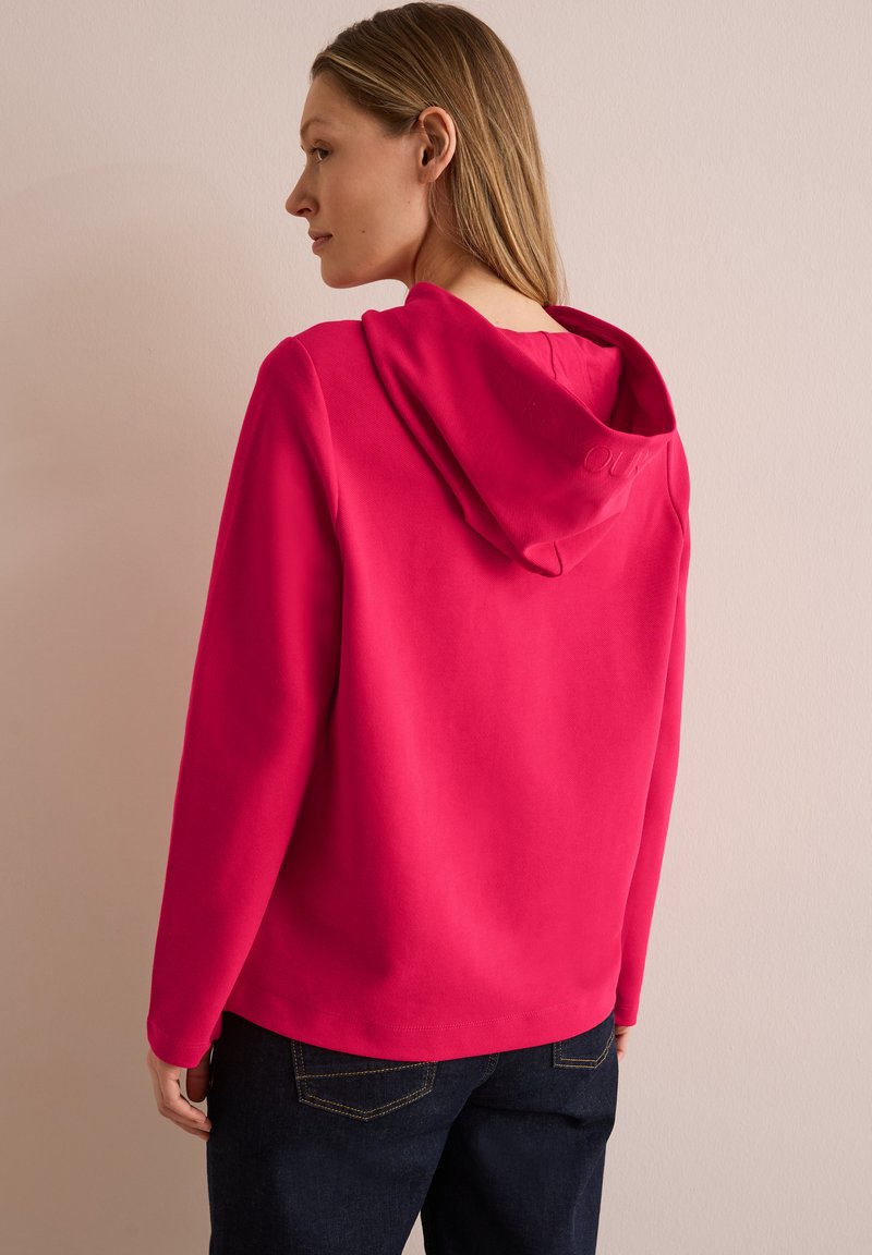 Sudadera fucsia con un corte relajado y un bolsillo canguro frontal, que presenta una capucha texturizada y puños acanalados. Combinada con unos jeans de mezclilla oscura.