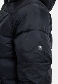 Närbild av en svart vadderad jackärm med en triangulär DC Shoe Co.-logotyp på överarmen.