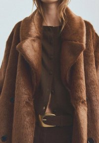 Brauner Kunstpelzmantel mit weitem Kragen und sichtbaren Knöpfen, kombiniert mit einem dunkelbraunen Oberteil und einem goldfarbenen Gürtelschnallen-Akzent.