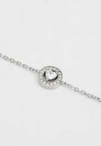 Silver bracelet featuring a circular centerpiece with a heart-shaped crystal surrounded by small clear stones, engraved with "Guess."