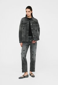 Schwarze Jeansjacke und passende Jeans mit Verzierungen, die am Saum leicht ausgefranst sind. Getragen mit flachen, gemusterten Schuhen.