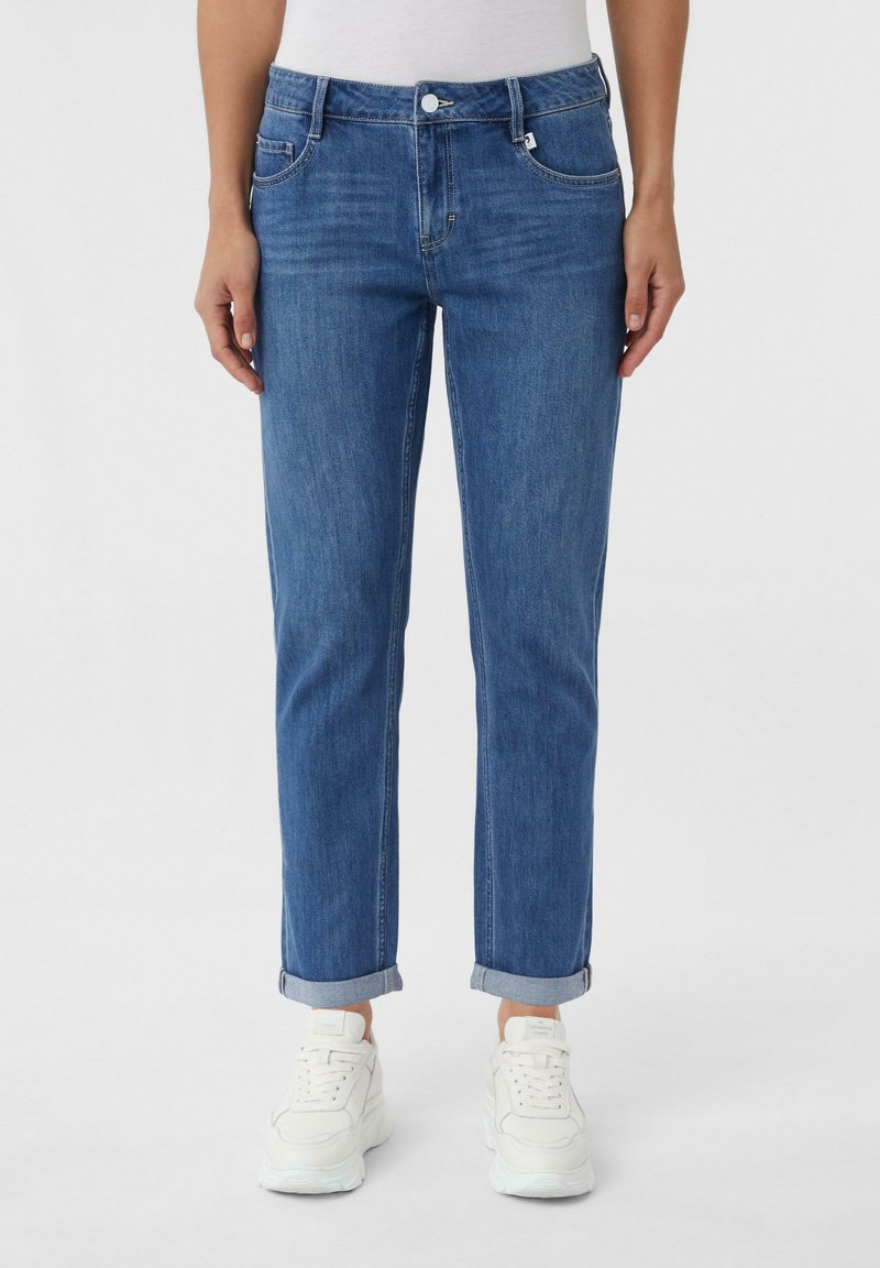 Niebieskie dżinsy o prostym kroju, z przednimi kieszeniami, podwiniętymi nogawkami i pojedynczym zapięciem na guzik. Połączone z białymi sneakersami.